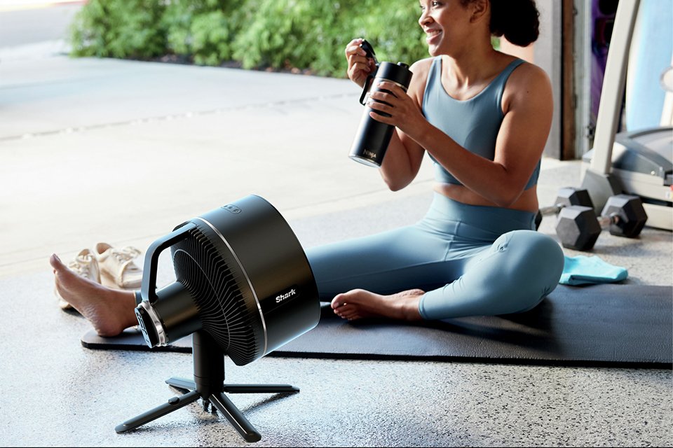 A woman sat in front of a Shark FlexBreeze Black Pedestal Fan on a yoga mat.